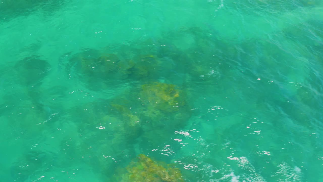 Turquoise ocean water at Shipwrecks Beach passes over dark submerged rocks and a yellow-green vegetation patch, with no visible people.