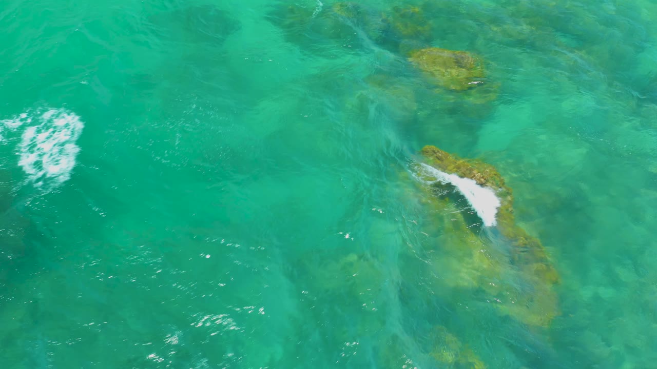 A small white foam patch appears over shallow turquoise water and darker submerged rocks at Shipwrecks Beach, Baja California Sur, with no visible people.