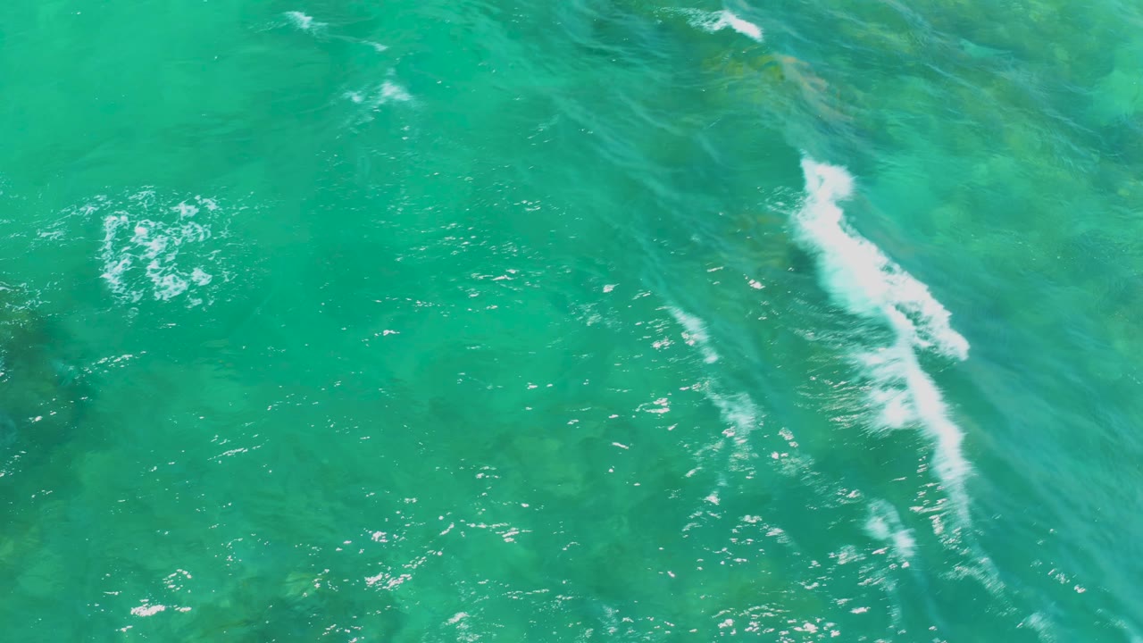 White wave foam crosses the edge of a submerged rock under bright turquoise water at Shipwrecks Beach, with no visible people.