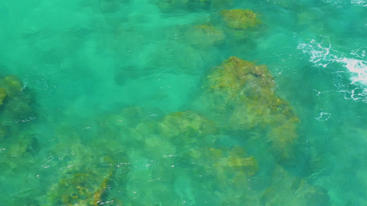 Aerial frame from Shipwrecks Beach showing green-blue water over submerged rocks, with a bright foam patch crossing near the center and no visible people.