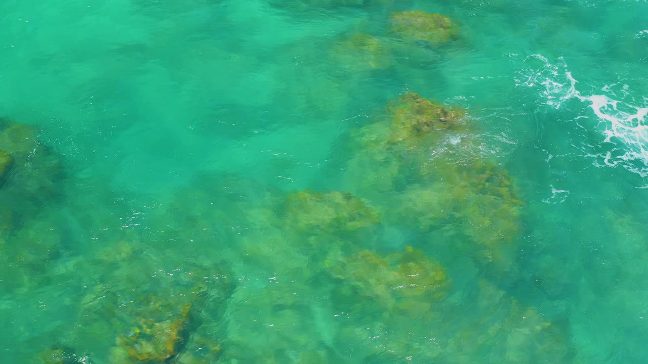 Overhead Shipwrecks Beach water view with scattered submerged rocks, a larger rock shape to the right, foam near the upper edge, and no visible people.