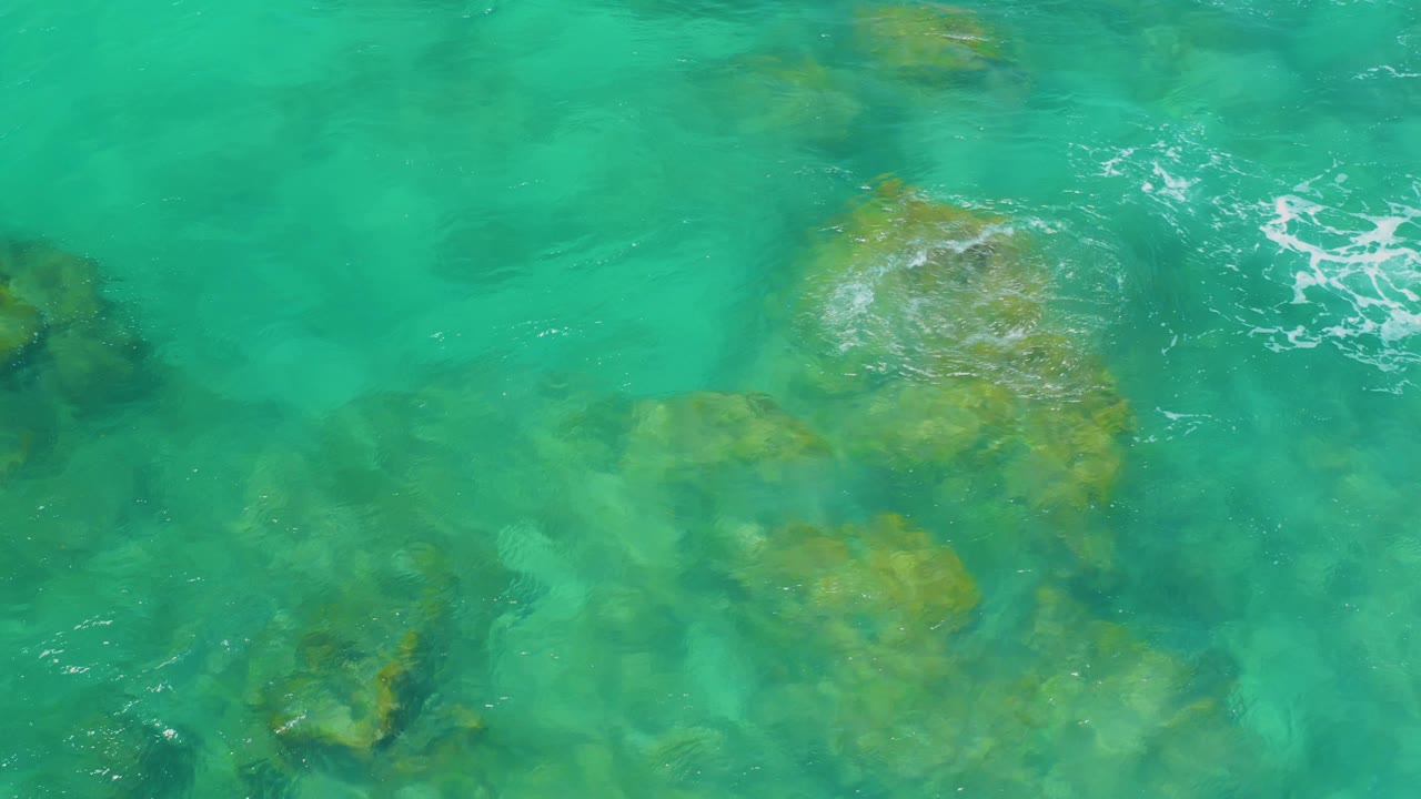 Teal water at Shipwrecks Beach covers a rounded submerged rock cluster, with a small white foam streak above it and no visible people.