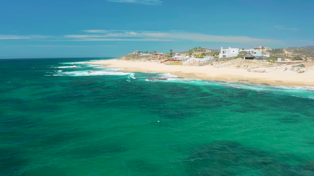 Opening aerial view at Shipwrecks Beach in Baja California Sur, with green ocean in the foreground, white surf on sand, low coastal buildings behind the beach, and no visible people.