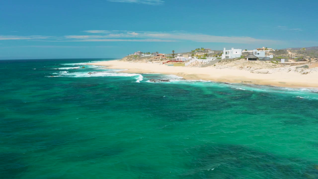 Turquoise water fills most of the frame at Shipwrecks Beach, with white surf tracing the sand, a row of low buildings on the dunes, and no visible people.