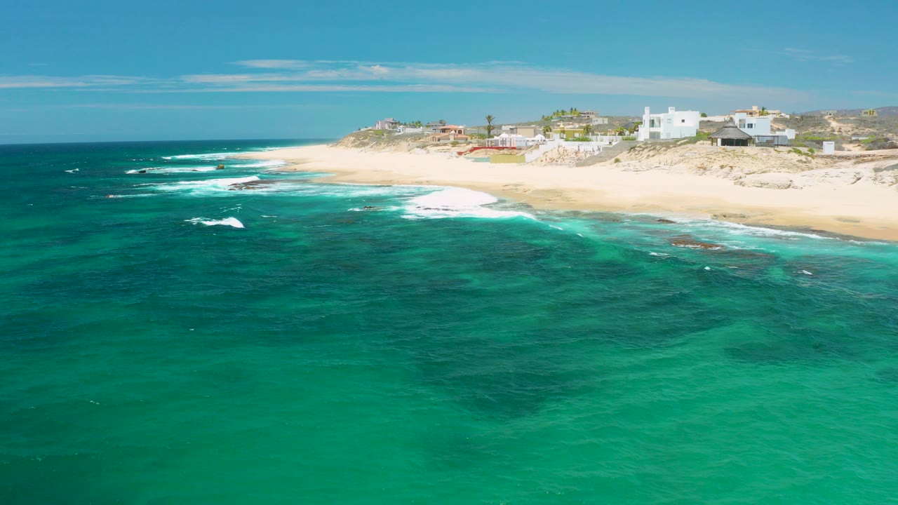 Opening aerial frame over Shipwrecks Beach in Baja California Sur, with turquoise water in the foreground, pale sand and low dune buildings beyond, and no visible people.