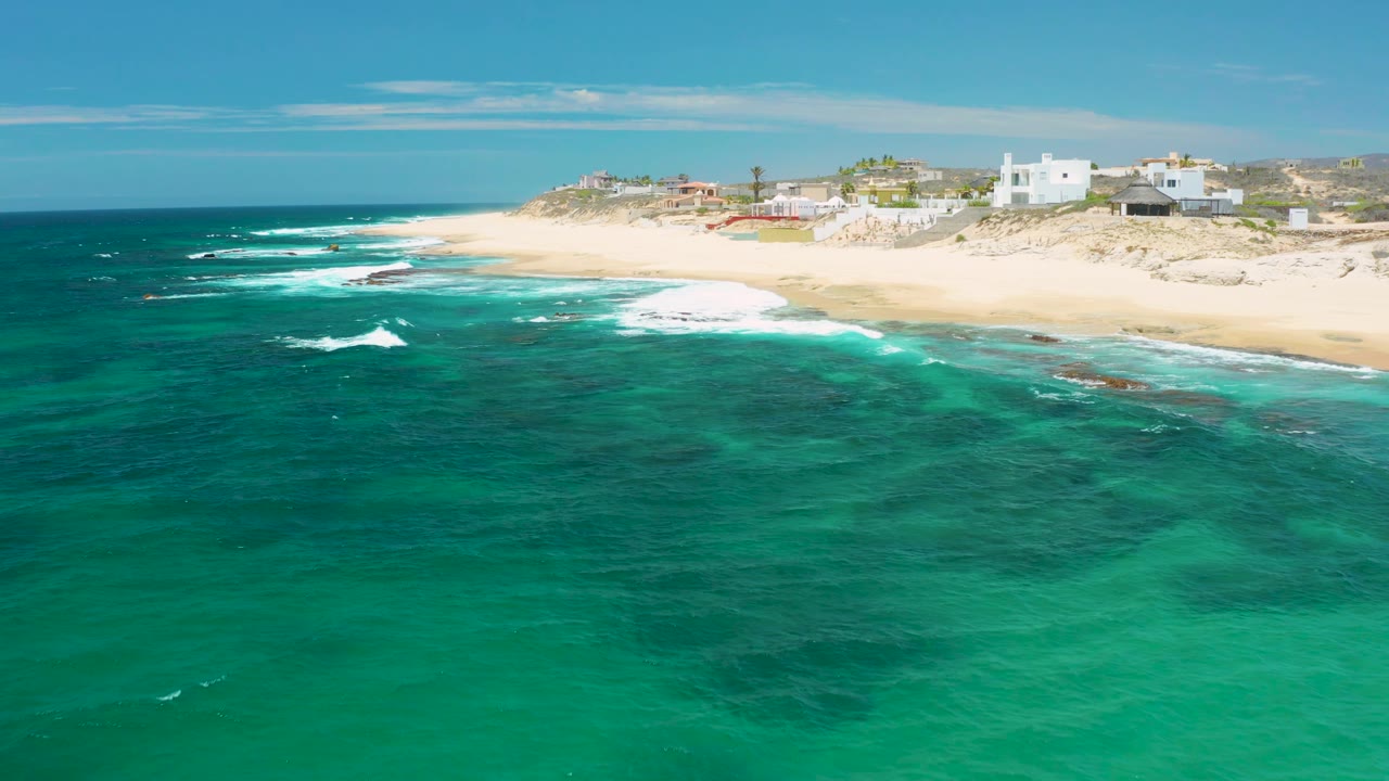 At 0:02, the frame holds a wide aerial view of Shipwrecks Beach, with darker underwater patches, small breaking waves, pale sand, dune vegetation, and no visible people.