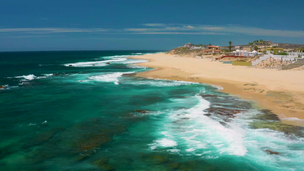 Aerial view of Shipwrecks Beach in Baja California Sur with turquoise surf breaking over dark rocks, tan sand to the right, low buildings on the bluff, and no visible people.