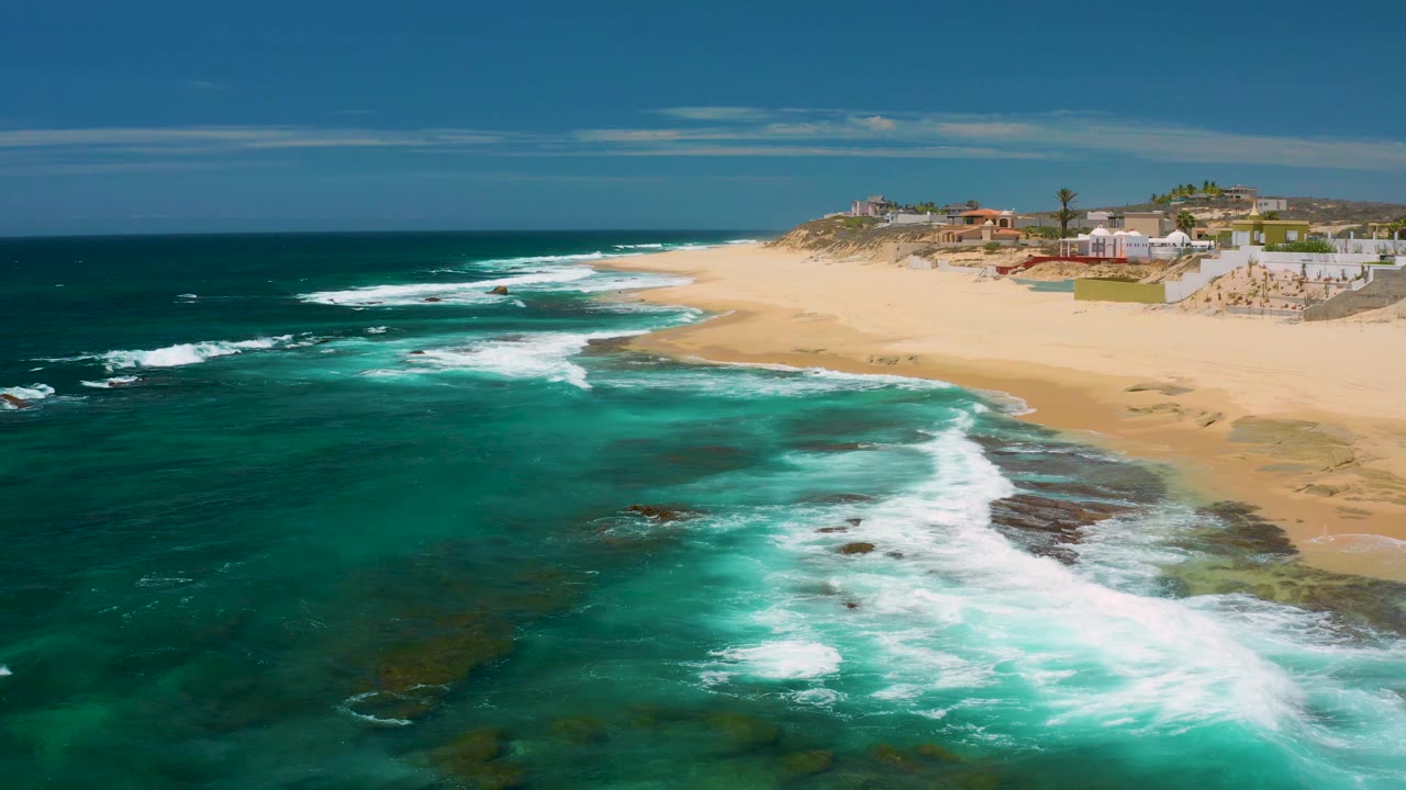 Aerial frame of Shipwrecks Beach with surf lines crossing the rocky shallows, a broad sand strip on the right, clustered low buildings beyond the beach, and no visible people.