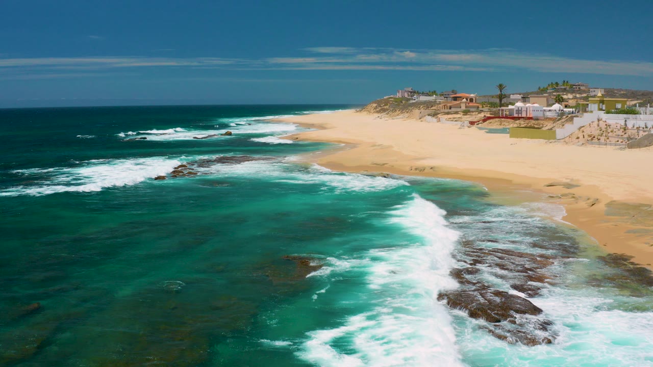 Opening aerial view at Shipwrecks Beach, Baja California Sur, with turquoise water, white surf over dark rock shelves, pale sand, distant beach buildings, and no visible people.
