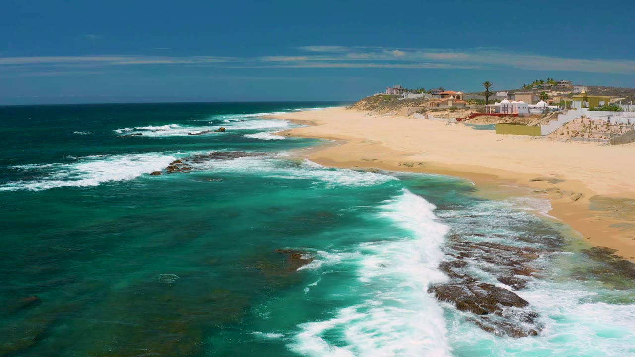 White waves roll toward a broad sandy beach with low buildings beyond it.