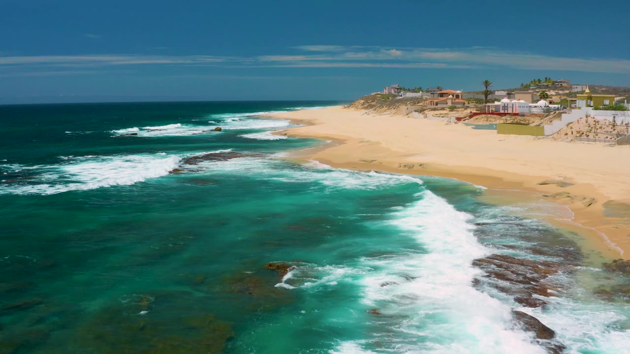 Aerial view over Shipwrecks Beach with foamy surf wrapping around exposed rocks, a broad empty-looking sand band, distant buildings, and no visible people.