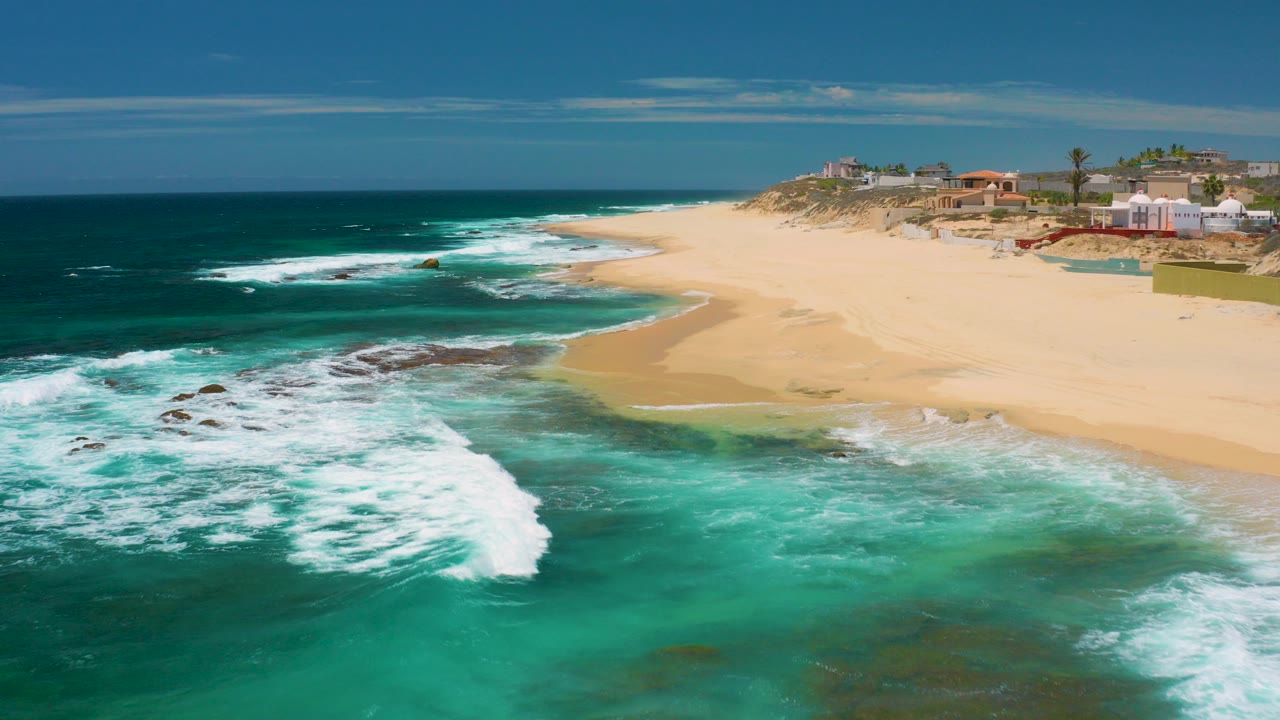 Aerial view at Shipwrecks Beach, Baja California Sur, with white surf spreading over dark rocks, pale sand curving inland, beachfront properties on the dune line, and no visible people.