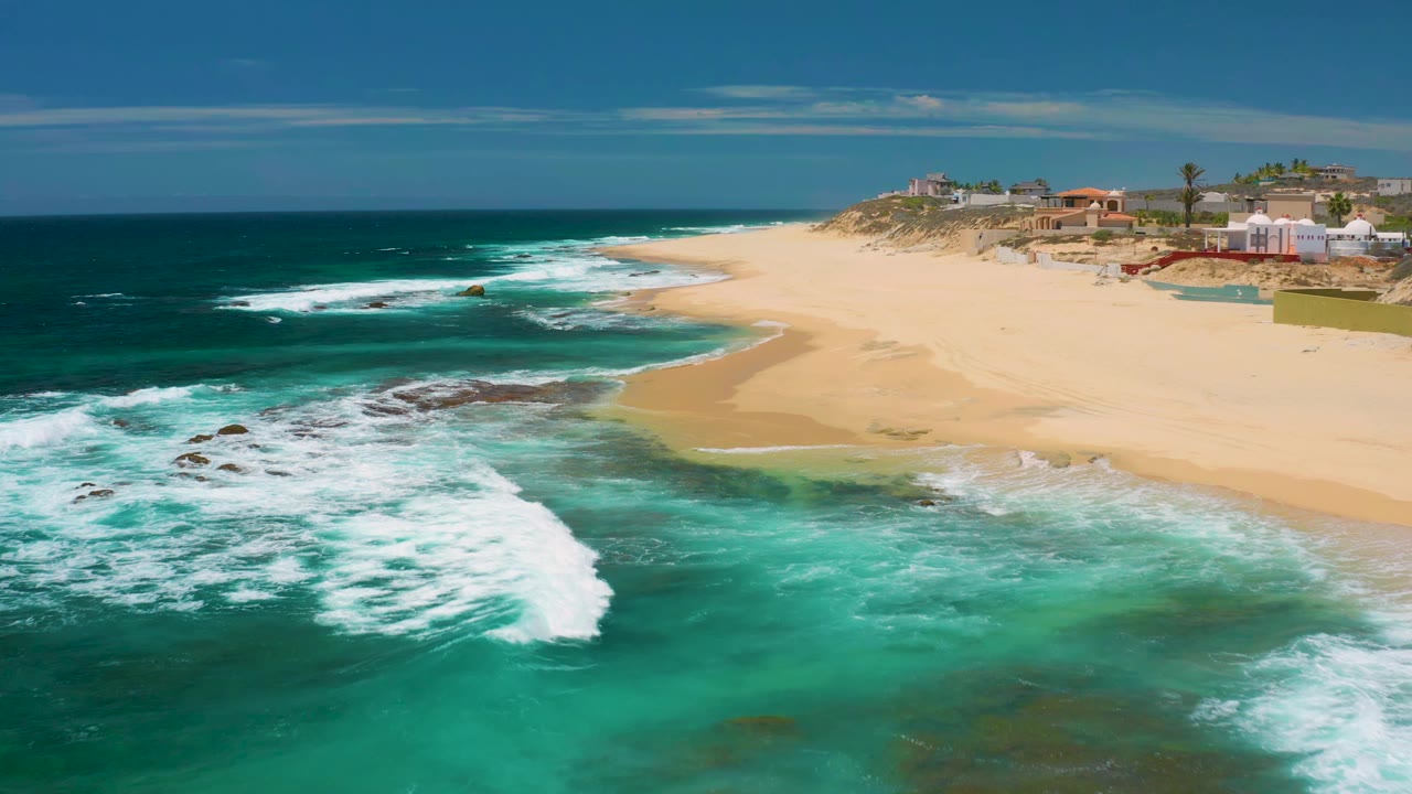 At 0:01, the aerial view holds on Shipwrecks Beach as a broad white wave pushes toward the sand, with rocky shallows in the foreground and no visible people on the beach.