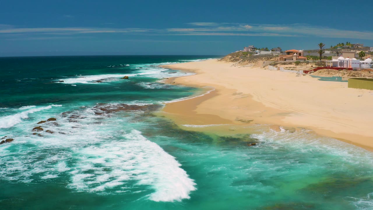 At 0:02, Shipwrecks Beach appears from above with foaming water crossing rock shelves, a wide band of empty sand, low coastal buildings beyond the dunes, and no visible people.