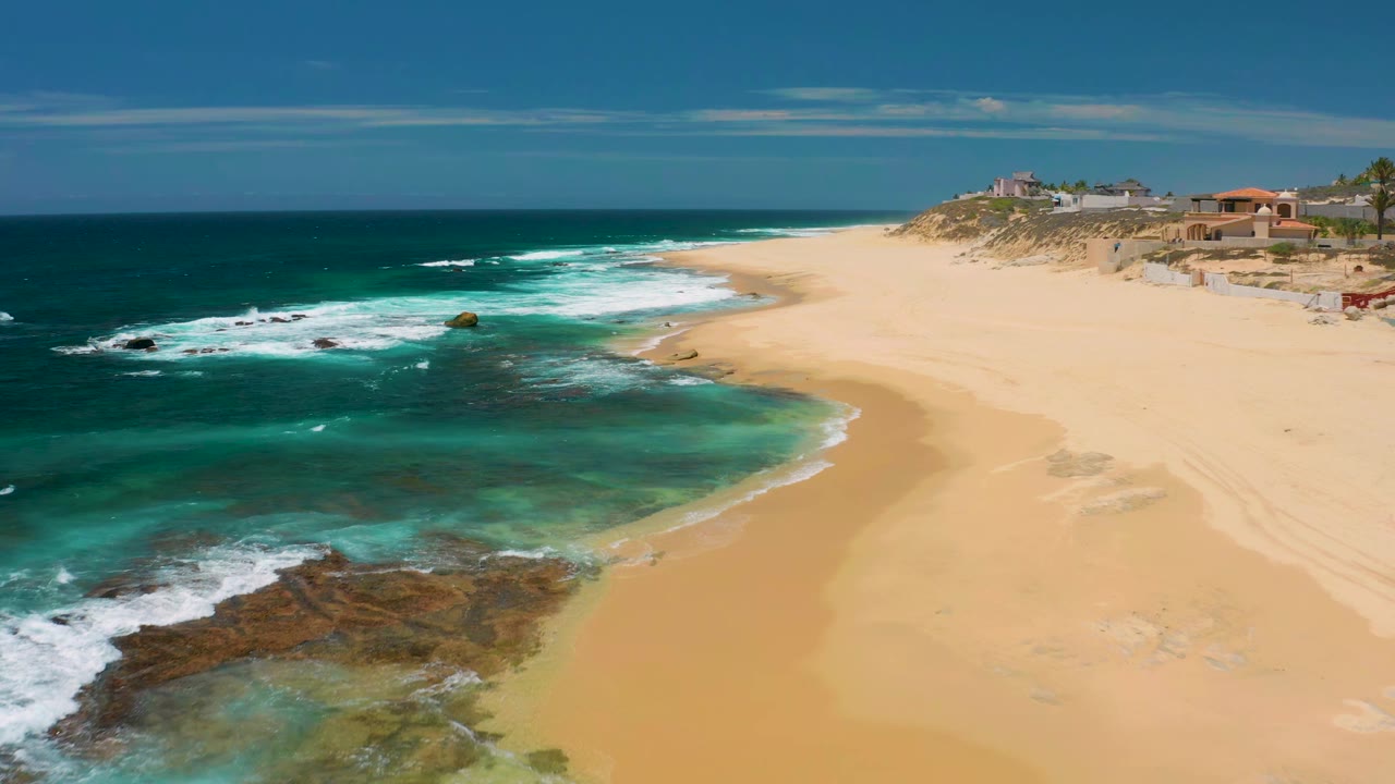 Aerial view at Shipwrecks Beach with white waves spreading over reef rock, clear green-blue shallows, curved wet sand, dune scrub, distant buildings, and no visible people.