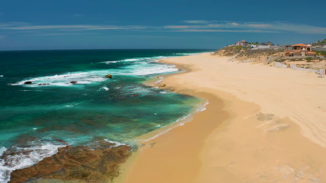 Shipwrecks Beach aerial frame with darker ocean on the left, foamy surf over rocks, a pale sand expanse to the right, low dune vegetation, distant buildings, and no visible people.