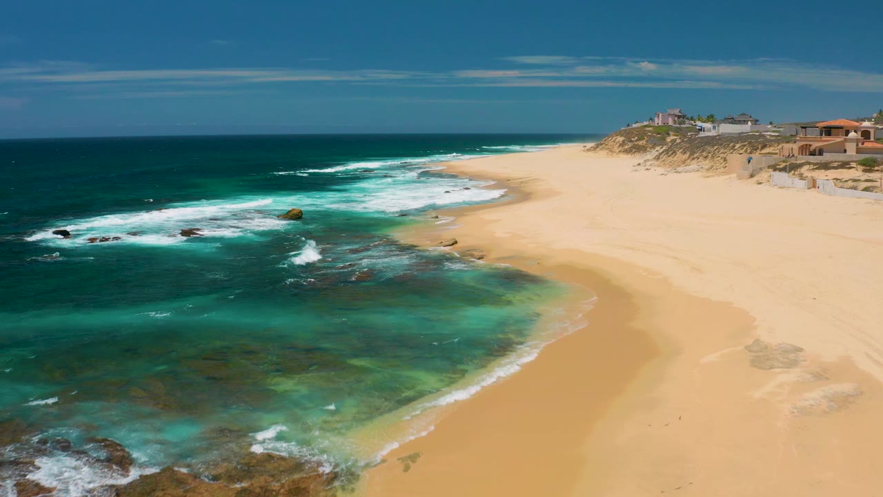 Wide coastal view of teal water, reef rocks, surf, beach, dunes, and bluff-top buildings.