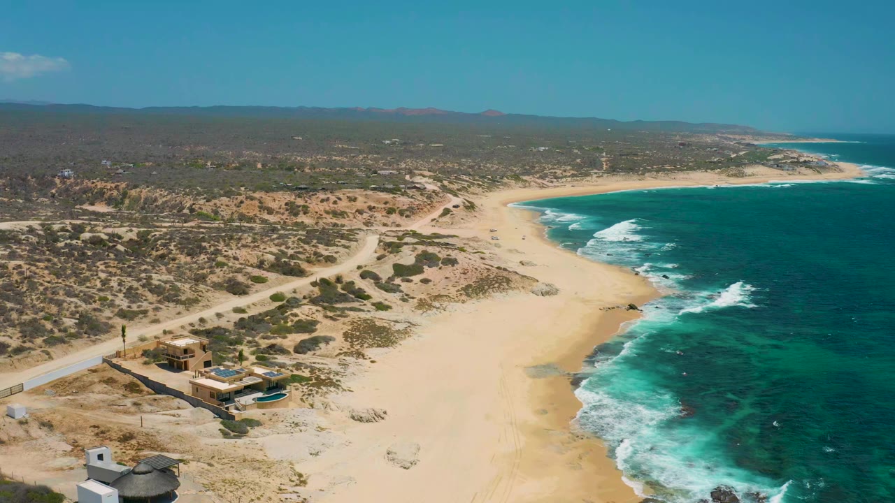 Aerial view over Shipwrecks Beach in Baja California Sur, Mexico, showing low buildings near the sand, dirt roads through scrub, turquoise surf on the right, and no visible people.