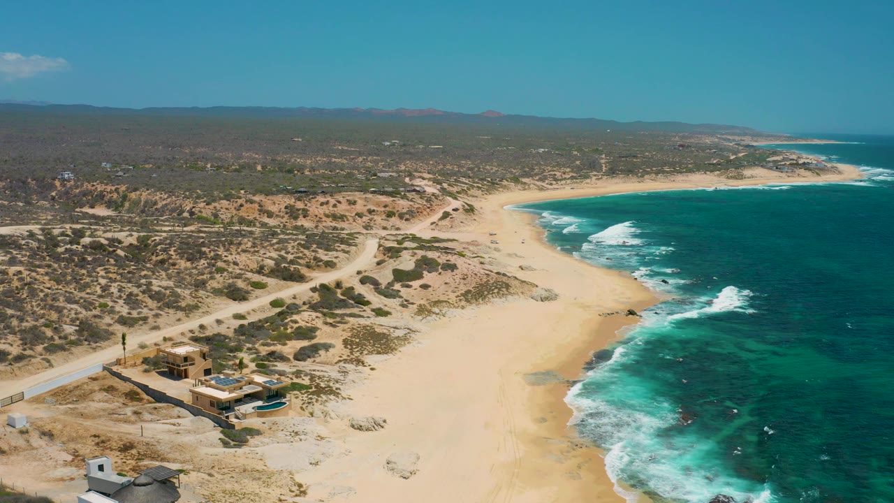 Drone-height view of Shipwrecks Beach showing more open sand in the center, turquoise water and white foam to the right, desert scrub and tracks to the left, and no visible people.