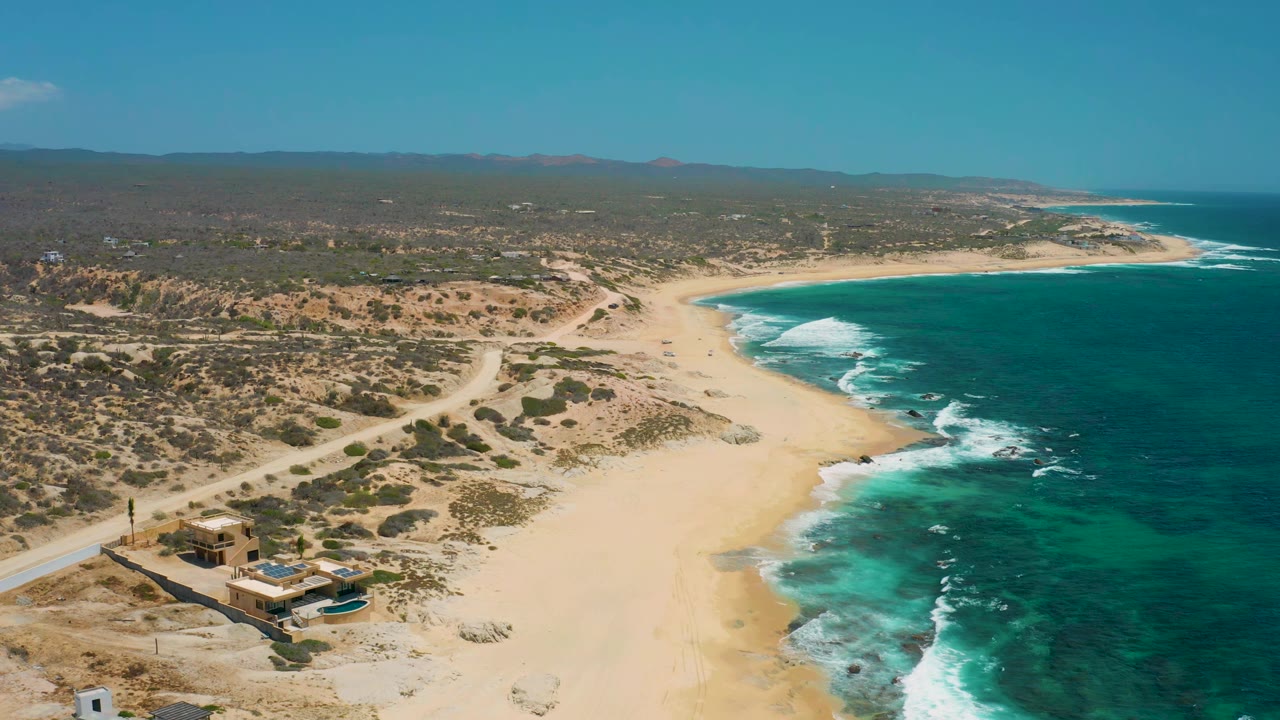 Wide aerial frame of Shipwrecks Beach showing the shoreline bending around blue-green water, scrubland behind the sand, a dirt road, a coastal building, and no visible people.
