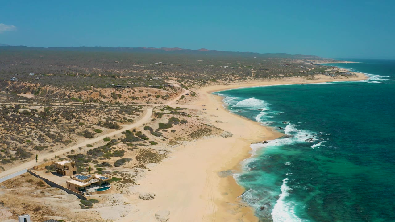 Aerial view at Shipwrecks Beach with empty sand in the foreground, white waves along the right edge, desert scrub inland, a beachfront structure near the lower left, and no visible people.