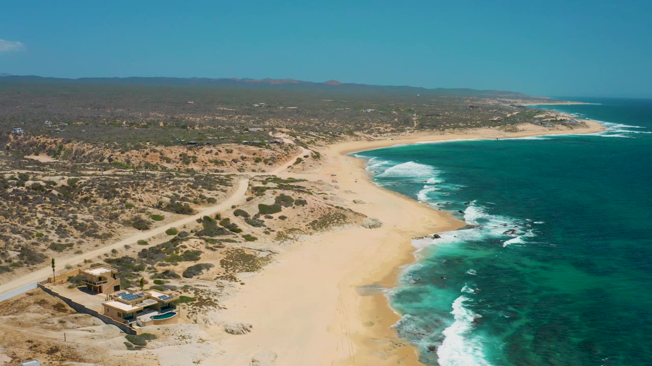 Drone view over a sandy coast with road tracks, surf line, shrubs, and a beach house.