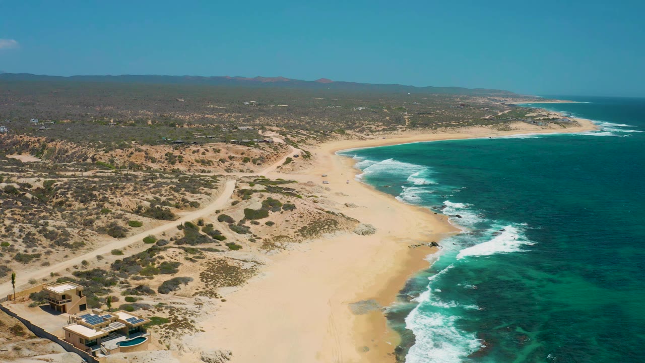A second aerial moment over Shipwrecks Beach shows the surf line slightly farther along the sand, coastal buildings at the lower left edge, inland dirt tracks, distant hills, and no visible people.