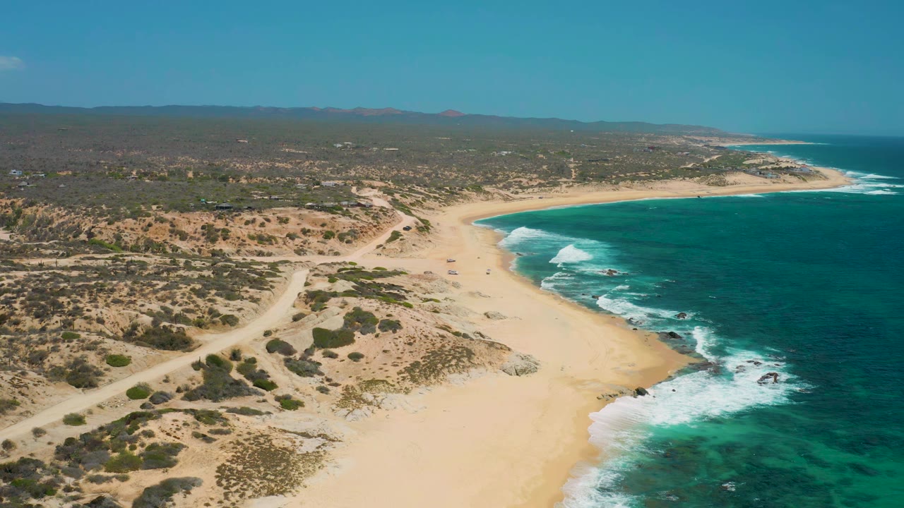 Aerial frame over Shipwrecks Beach in Baja California Sur, showing the curved sandy bay, turquoise shallows, darker offshore water, inland scrub, and no visible people.