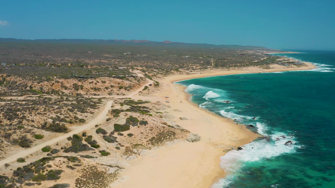 Wide drone-style view of Shipwrecks Beach in Baja California Sur, with surf breaking around rocks near shore, broad sand, low scrub inland, and no visible people.
