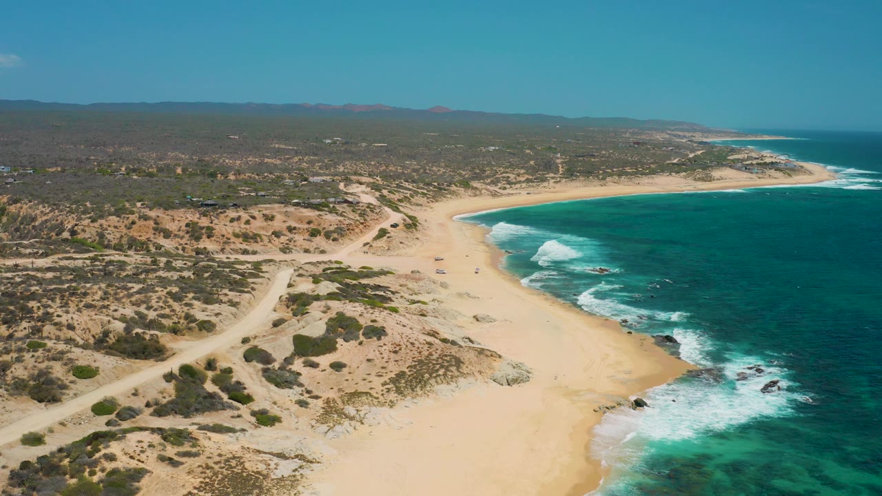 Aerial view along Shipwrecks Beach in Baja California Sur, with dirt tracks crossing scrubby sand, a broad empty beach, whitewater at the edge, and no visible people.