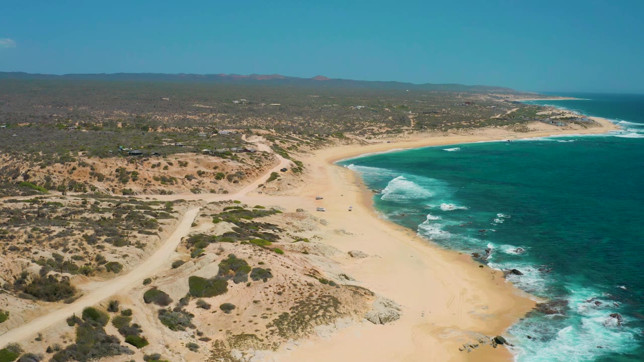 Wide drone view of curving beach with waves and desert tracks