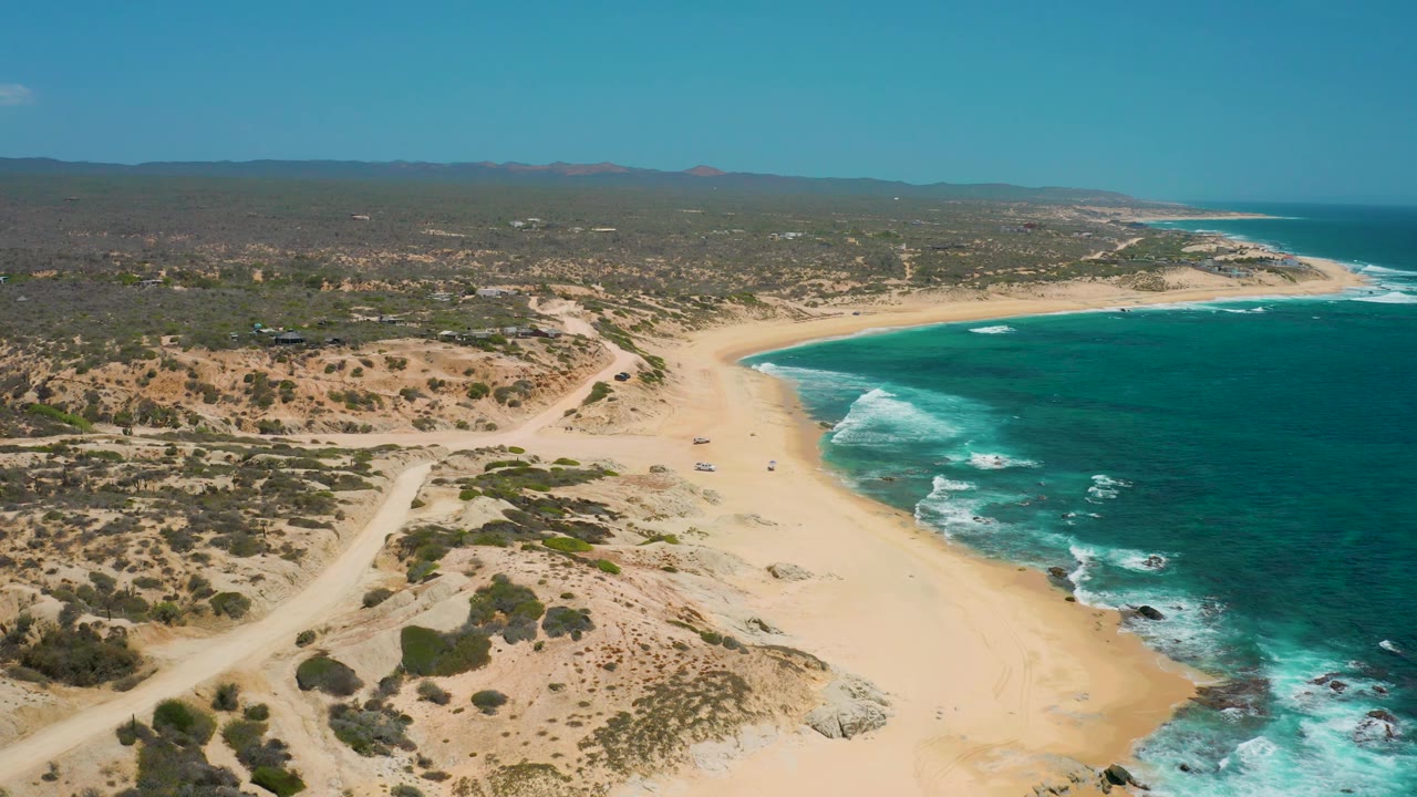 High view of Shipwrecks Beach with a sandy cove, turquoise sea on the right, white foam over rocks, scrubby inland terrain, dirt paths, and tiny people on the sand.