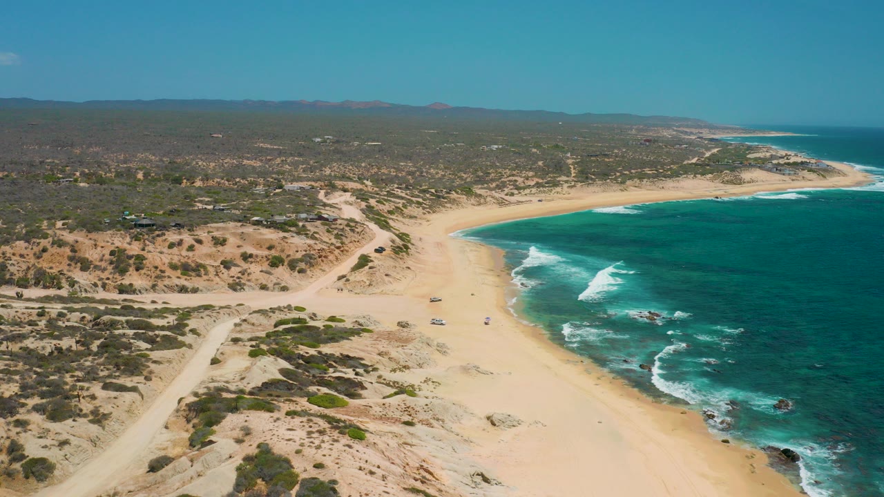 Aerial frame over Shipwrecks Beach in Baja California Sur, Mexico, showing pale sand curving beside turquoise water, scrub-covered dunes, dirt tracks, distant hills, and no visible people.
