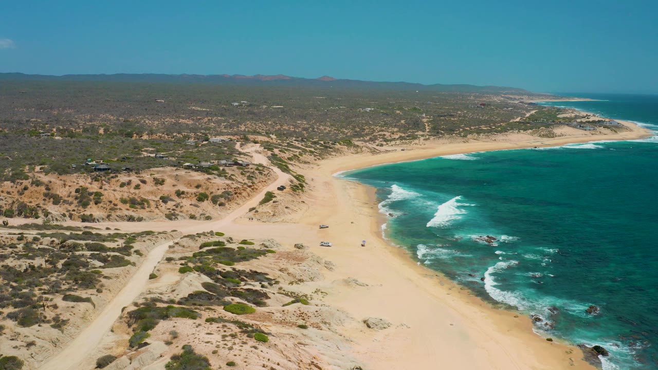 The coastline at Shipwrecks Beach fills the frame with open sand, a turquoise cove-like bend, white surf over nearshore rocks, scrubby inland ground, distant hills, and no visible people.