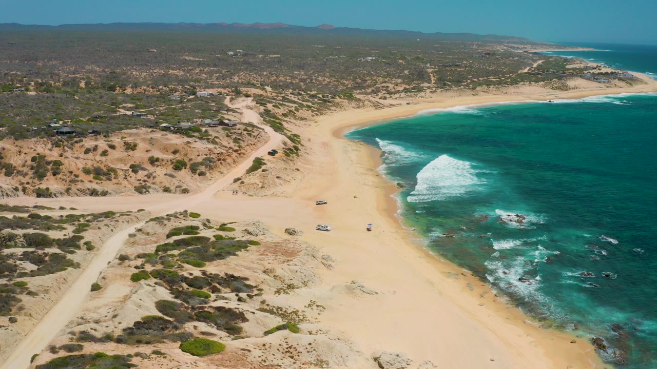 Opening aerial frame over Shipwrecks Beach in Baja California Sur, with pale sand curving beside turquoise water, scrub dunes, dirt access tracks, parked vehicles, and distant low buildings.