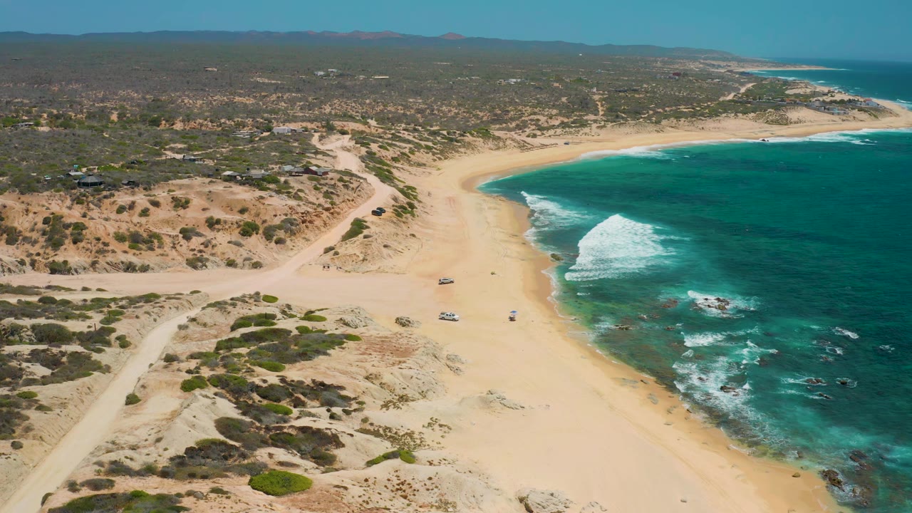 Aerial coastline view with surf breaking near a sandy beach and dirt roads through dry vegetation.