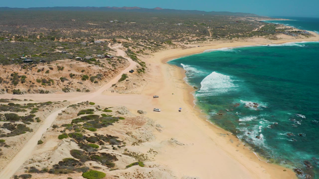 At 0:02, the aerial view holds a broad Shipwrecks Beach cove, with a larger white wave rolling toward shore and scattered vehicles set back on the sand.