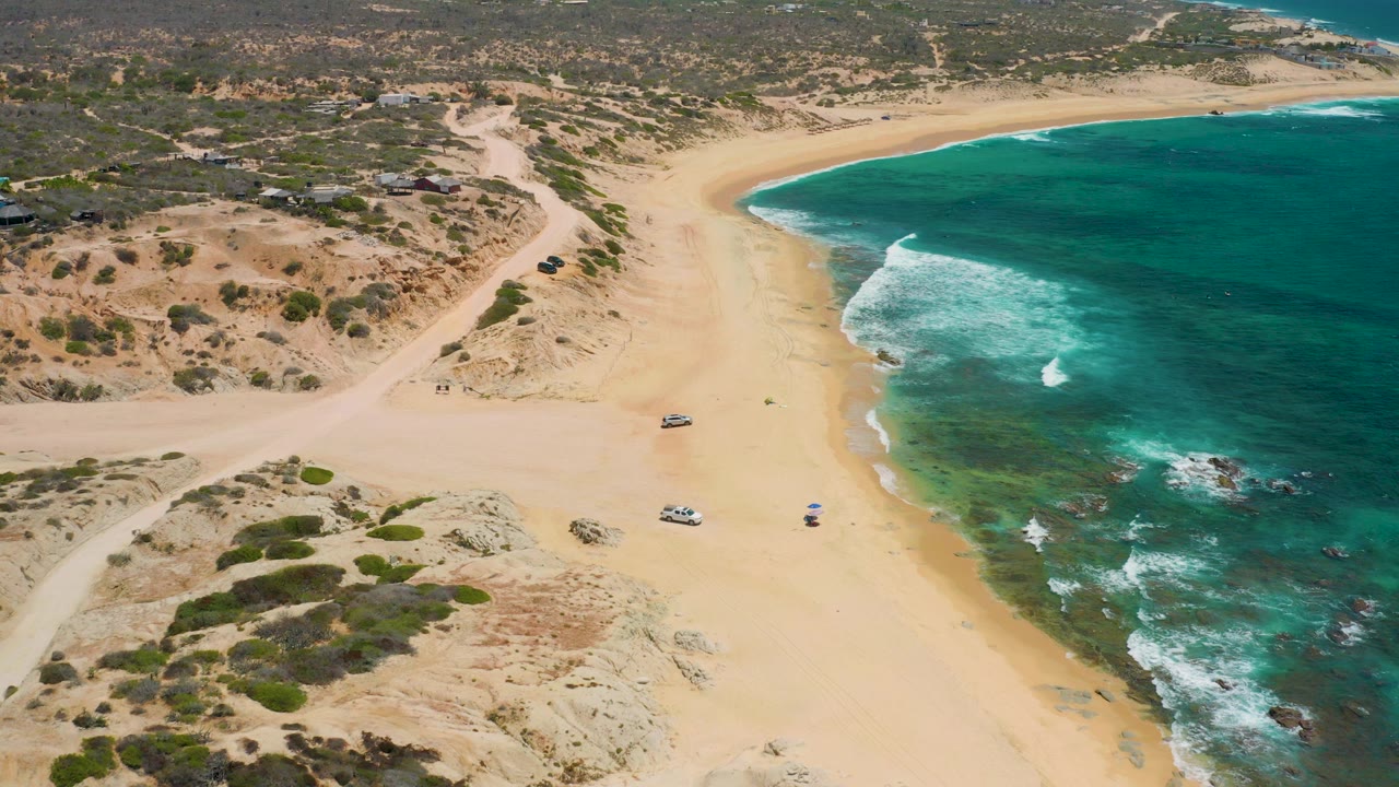 Opening aerial frame over Shipwrecks Beach in Baja California Sur, with pale sand, turquoise surf, scrub dunes, dirt tracks, parked vehicles, and tiny beach activity near the waterline.