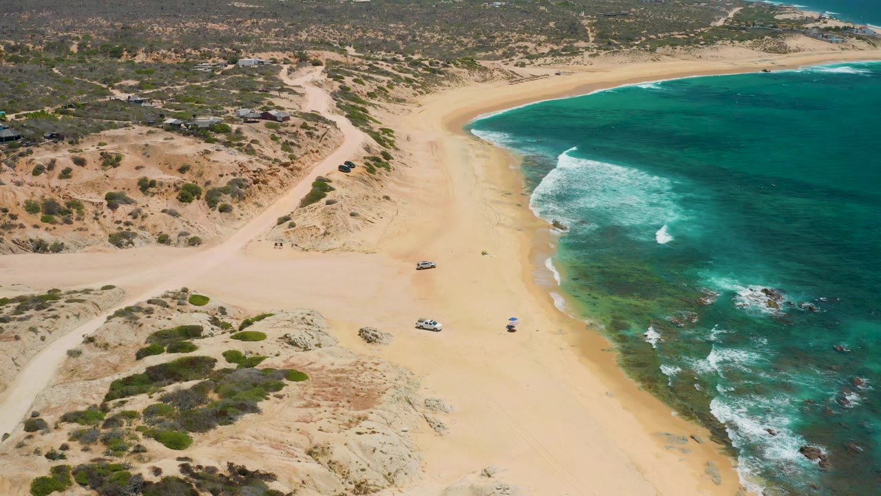 High view of curved beach with surf, scrub dunes, dirt tracks, and parked vehicles on the sand.