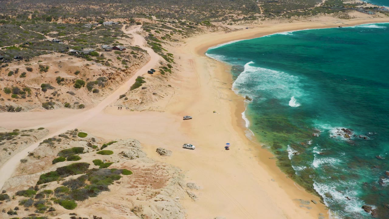 Shipwrecks Beach appears from above with ocean taking the right half of frame, sand and scrubland on the left, and a few vehicles spaced along the beach access area.
