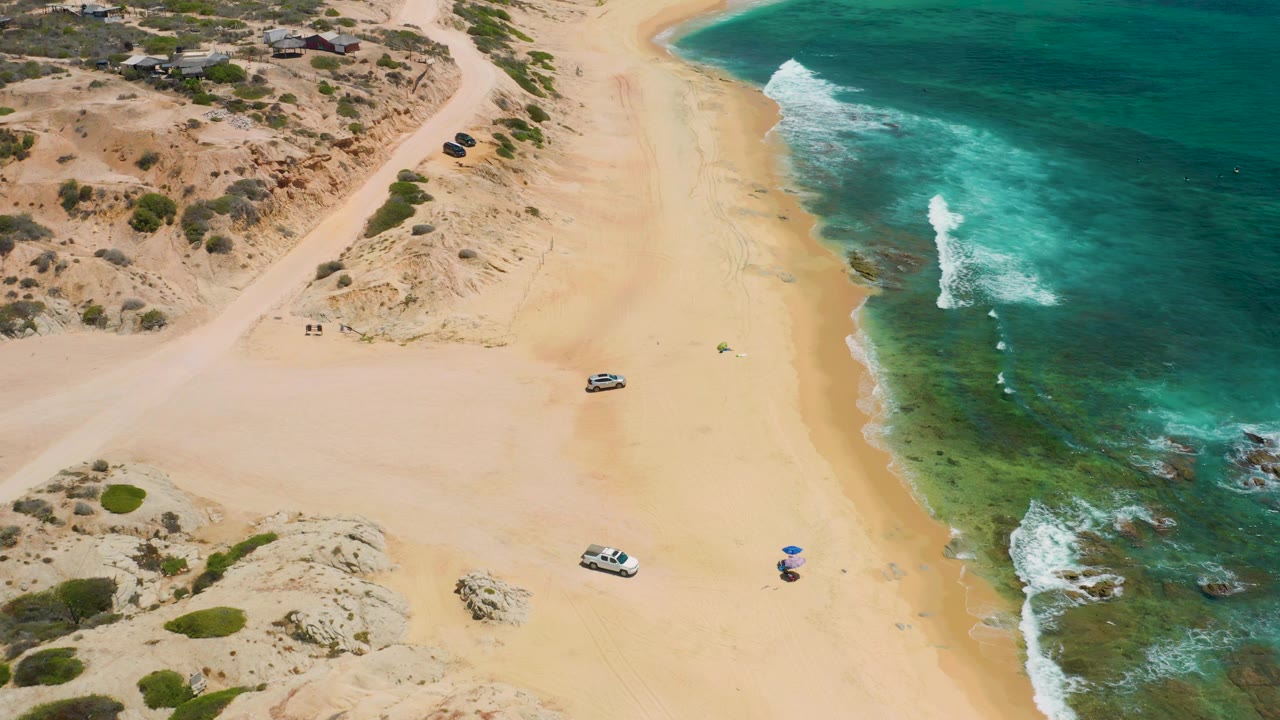 High aerial frame at Shipwrecks Beach showing the shoreline angled through the image, with clear green-blue shallows, light sand, dune scrub, parked vehicles, umbrellas, and no visible people.