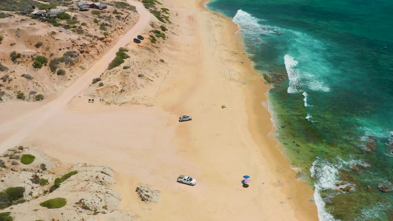 Aerial view of Shipwrecks Beach with a broad empty sand area, rocky green shallows, white wave lines, dunes and scrub at the top, a white vehicle on the beach, and no visible people.