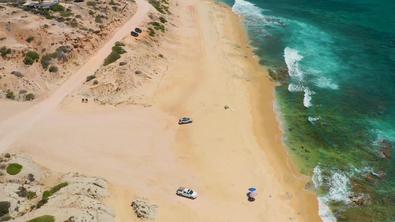 Drone-style aerial frame over Shipwrecks Beach with a white vehicle near the lower sand, two small umbrellas near the surf side, low dunes to the left, green water to the right, and no visible people.