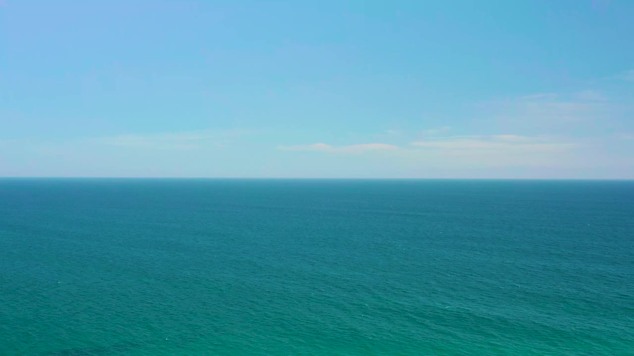 Wide aerial view over turquoise ocean at Shipwrecks Beach, Baja California Sur, with a straight horizon, pale clouds, and no visible people.