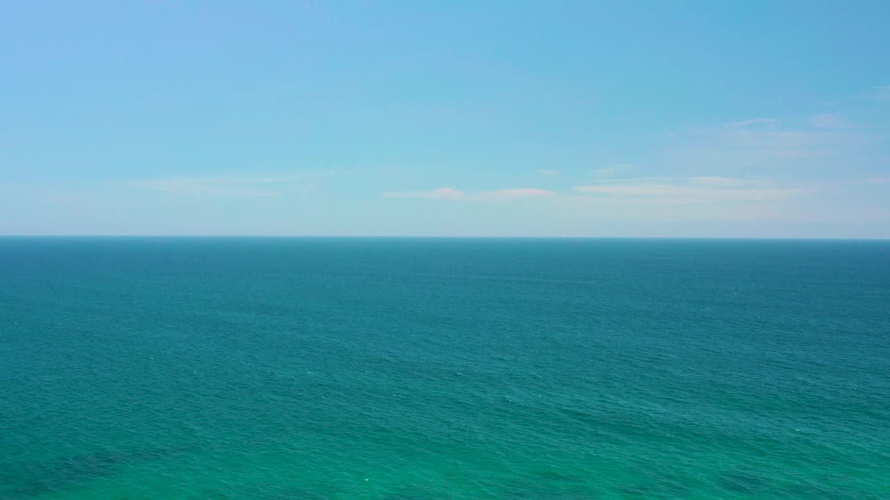 Mid-clip aerial frame of Shipwrecks Beach water showing turquoise surface texture, darker offshore bands, a level horizon, thin clouds, and no visible people.