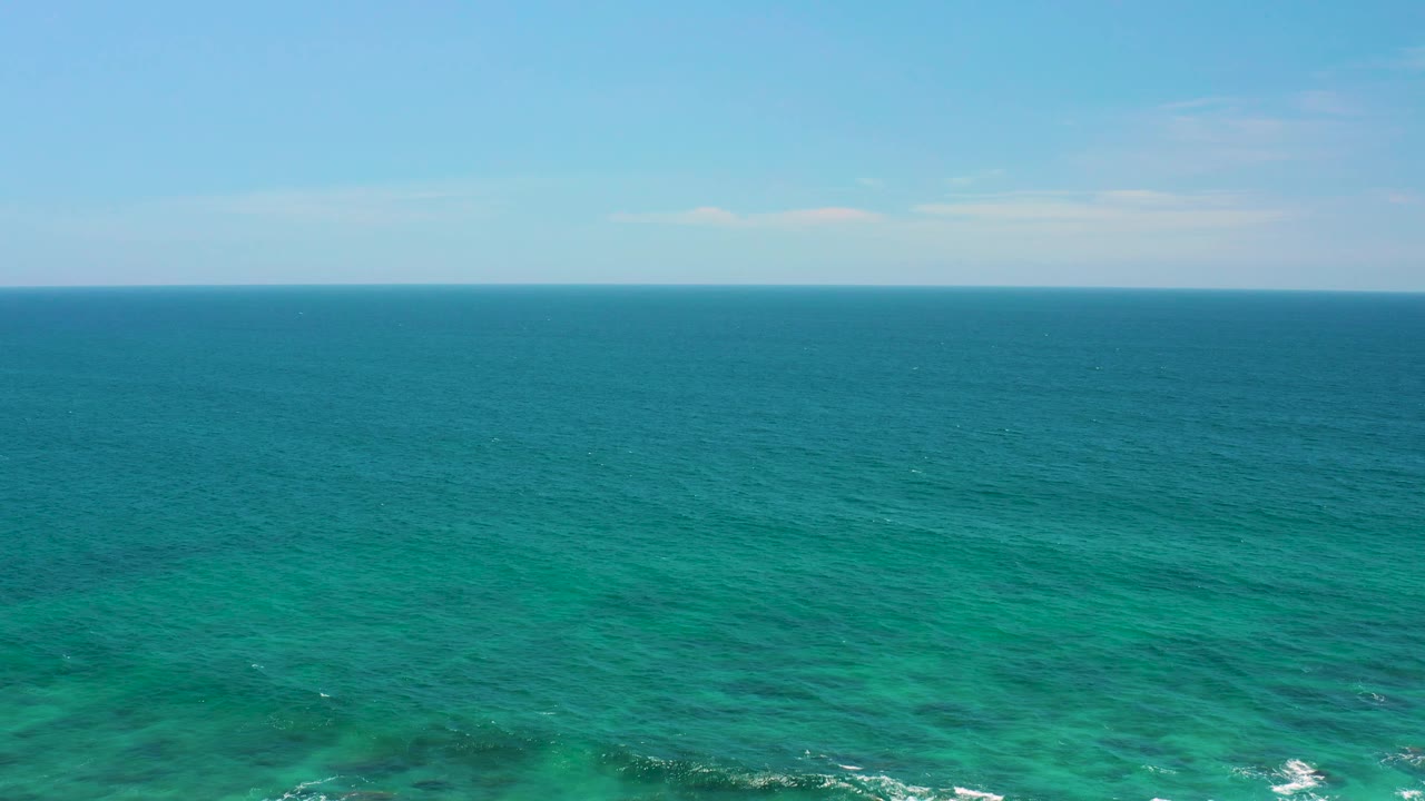 Aerial-looking frame at Shipwrecks Beach with turquoise water, darker underwater patches near the lower frame, a level horizon, and no visible people.
