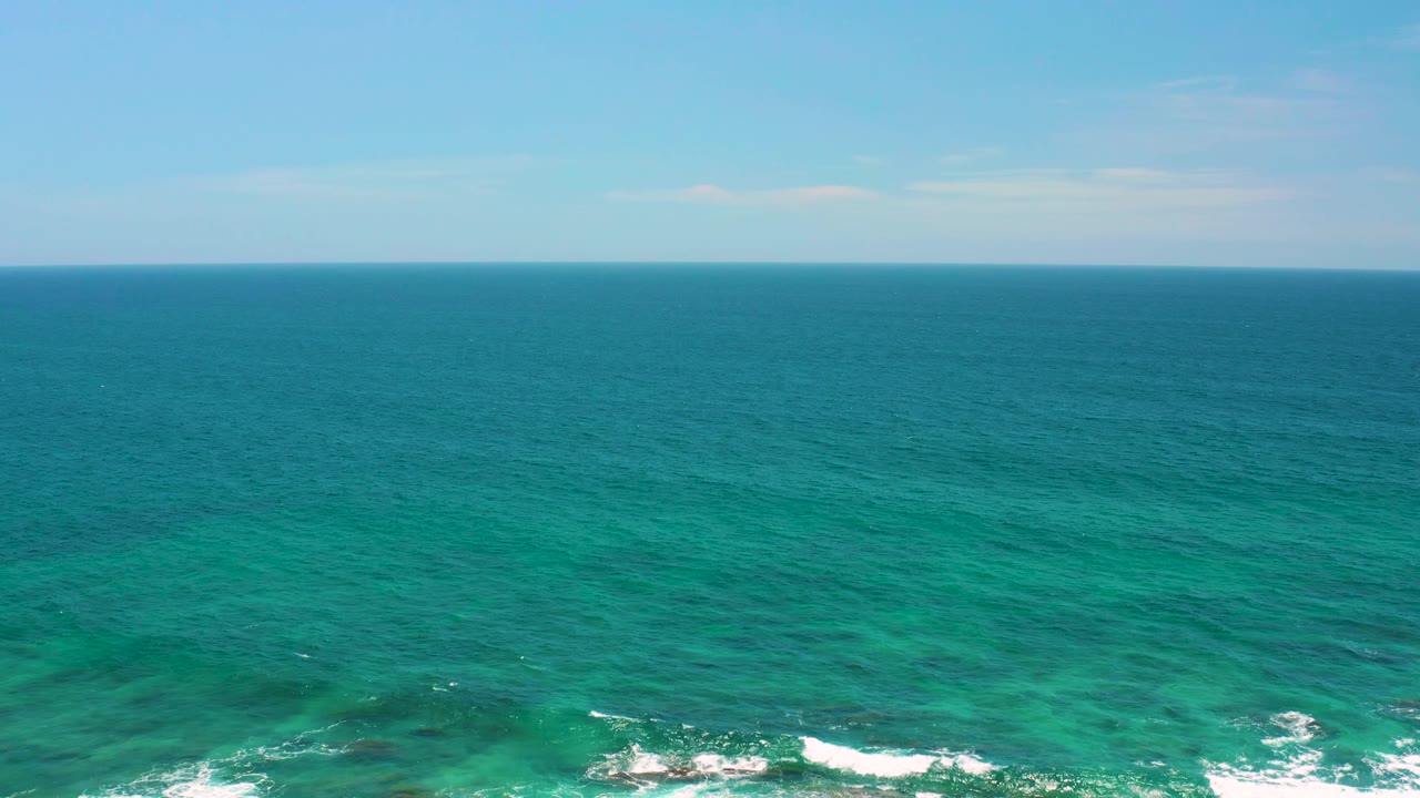 Mid-clip view of Shipwrecks Beach water, with open blue ocean stretching to a straight horizon, pale clouds above, and no visible people.