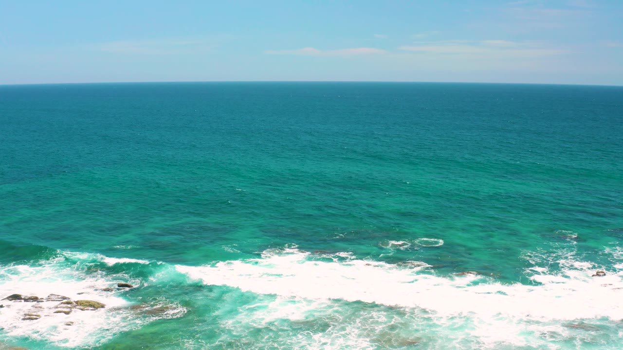 White surf breaks in a broad band beneath a clear ocean horizon at Shipwrecks Beach, Baja California Sur, with scattered rocks visible in the foam and no visible people.