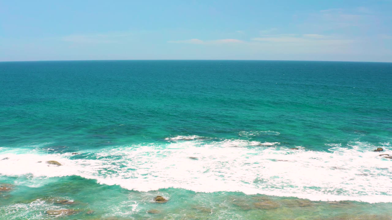 A later moment at Shipwrecks Beach shows whitewater spreading wider across the shallow rocky foreground, with open turquoise sea and no visible people.