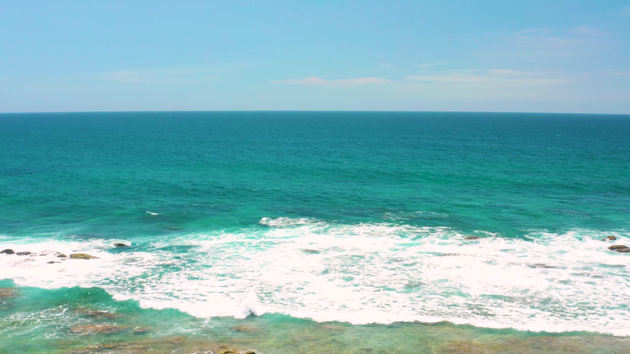 Mid-clip view of Shipwrecks Beach with churning white surf around exposed rocks, turquoise water stretching to the horizon, and no visible people.
